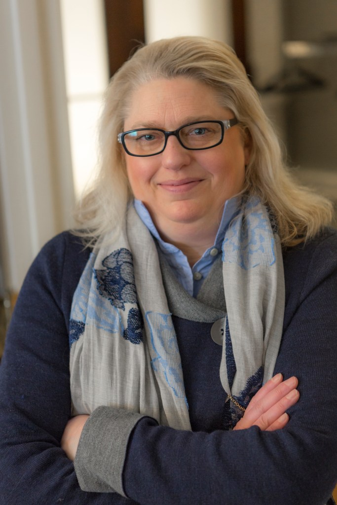 Image of Stacy Doore. 
Woman with blonde hair and blue eyes wearing glasses. She has a navy sweater, blue shirt and a gray and blue floral scarf. This is a professional looking image.